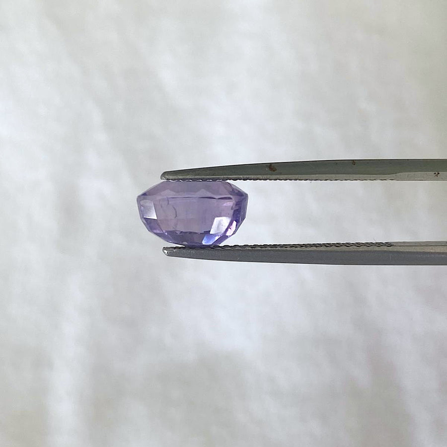 A faceted purple sapphire held between metal tweezers against a soft white textured background.