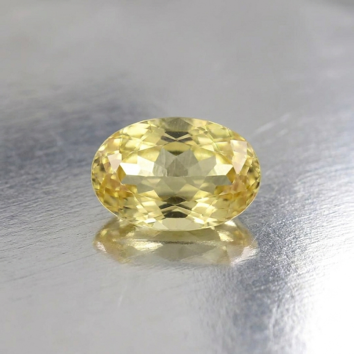 An oval-cut yellow gemstone with faceted reflections, resting on a reflective silver surface.