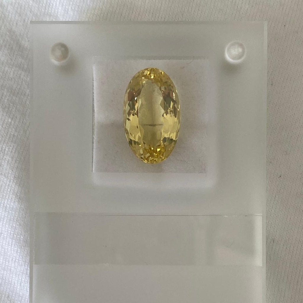 A faceted oval yellow gemstone displayed on a translucent white acrylic holder against a white fabric background.
