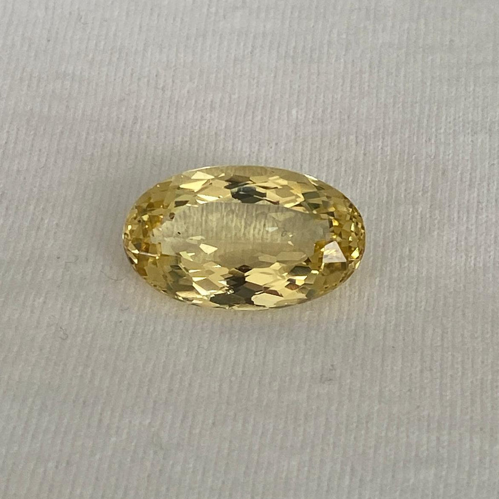 An oval-cut yellow gemstone with a faceted cut, displayed on a textured off-white background.