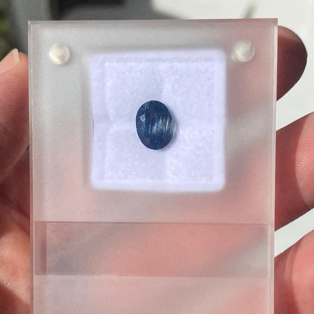 A hand holding a translucent acrylic display card with a small oval dark blue gemstone mounted on a white backing insert.