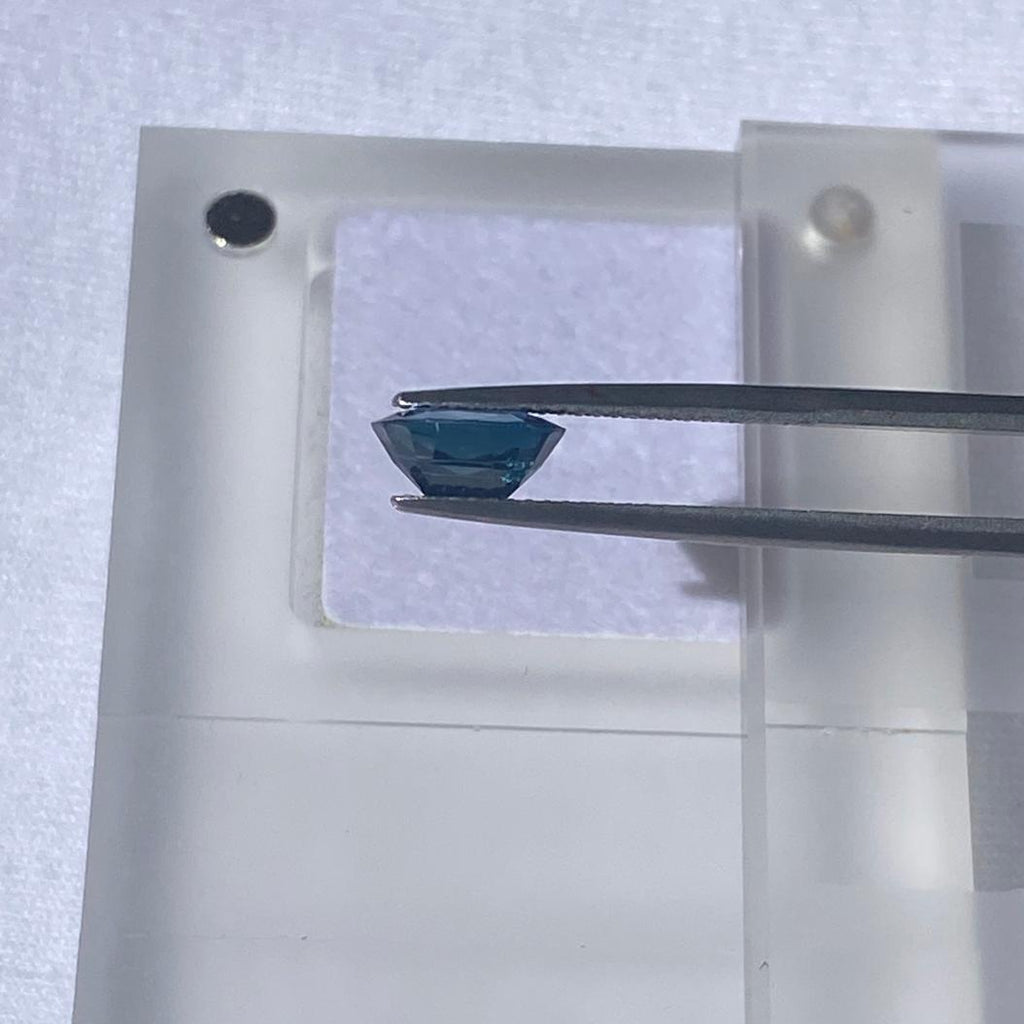 A faceted blue sapphire held by metal tweezers over a translucent acrylic display block on a light fabric background.