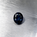 An oval-cut dark blue sapphire, displayed on a textured silver metallic surface.