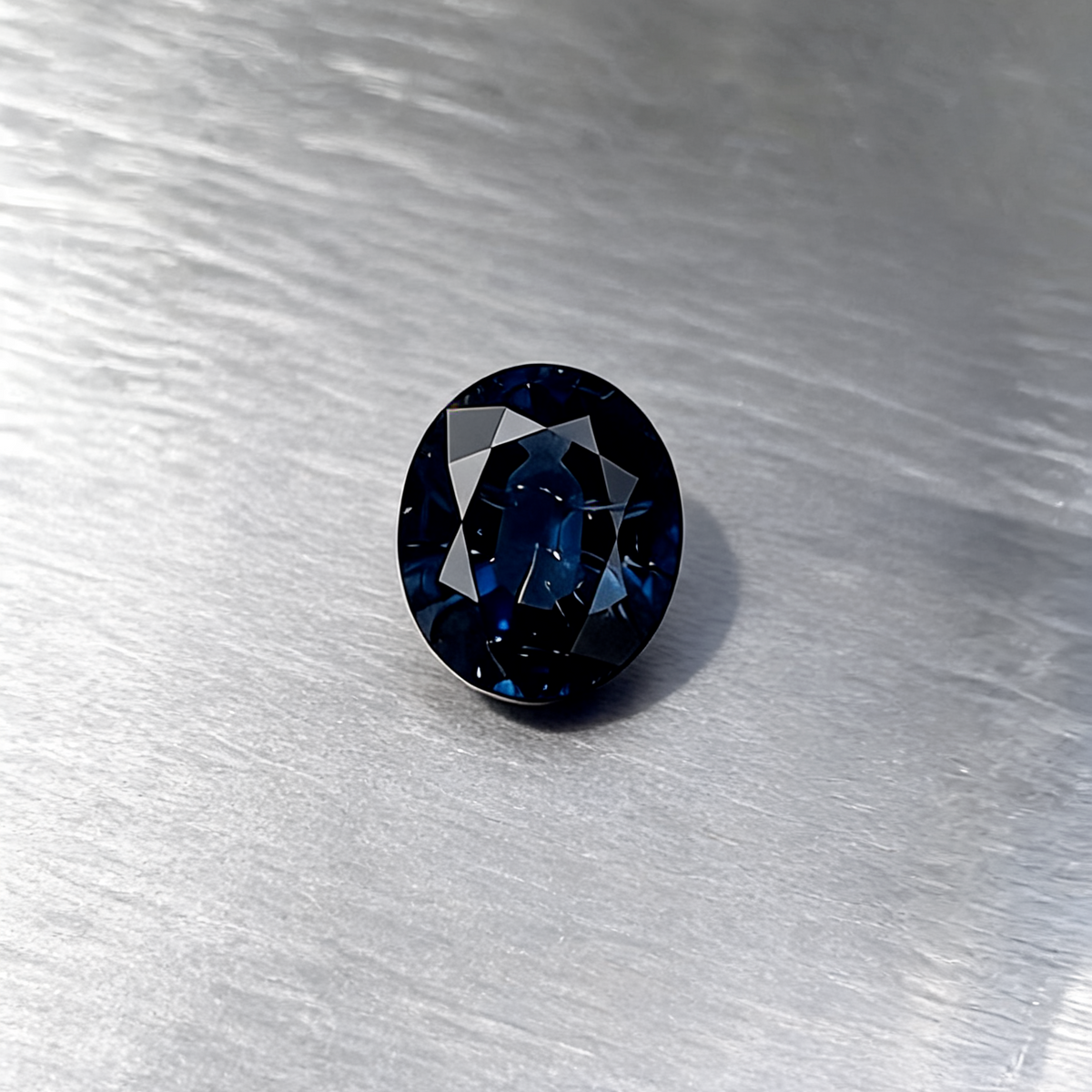 An oval-cut dark blue sapphire, displayed on a textured silver metallic surface.