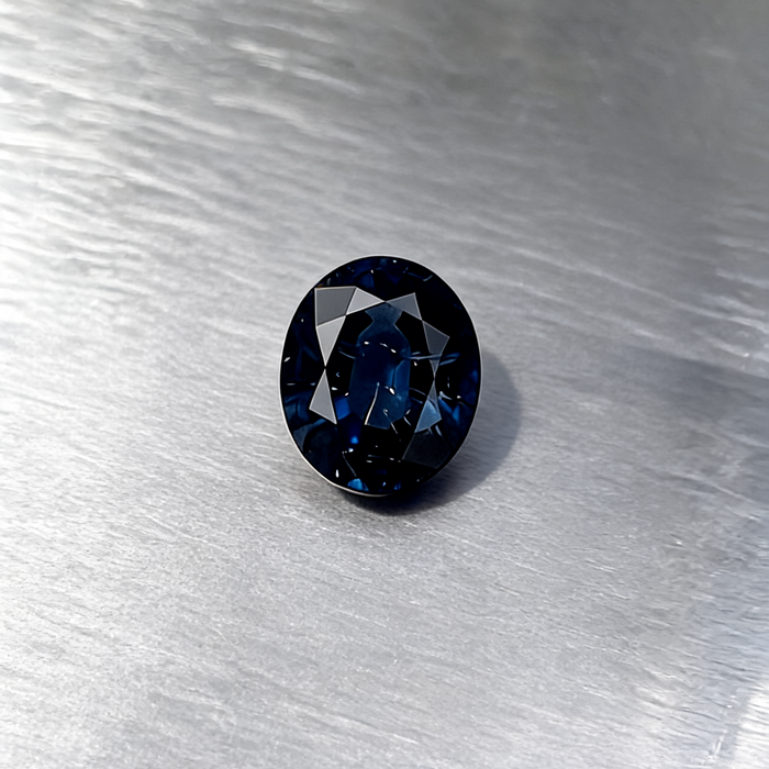 An oval-cut dark blue sapphire, displayed on a textured silver metallic surface.