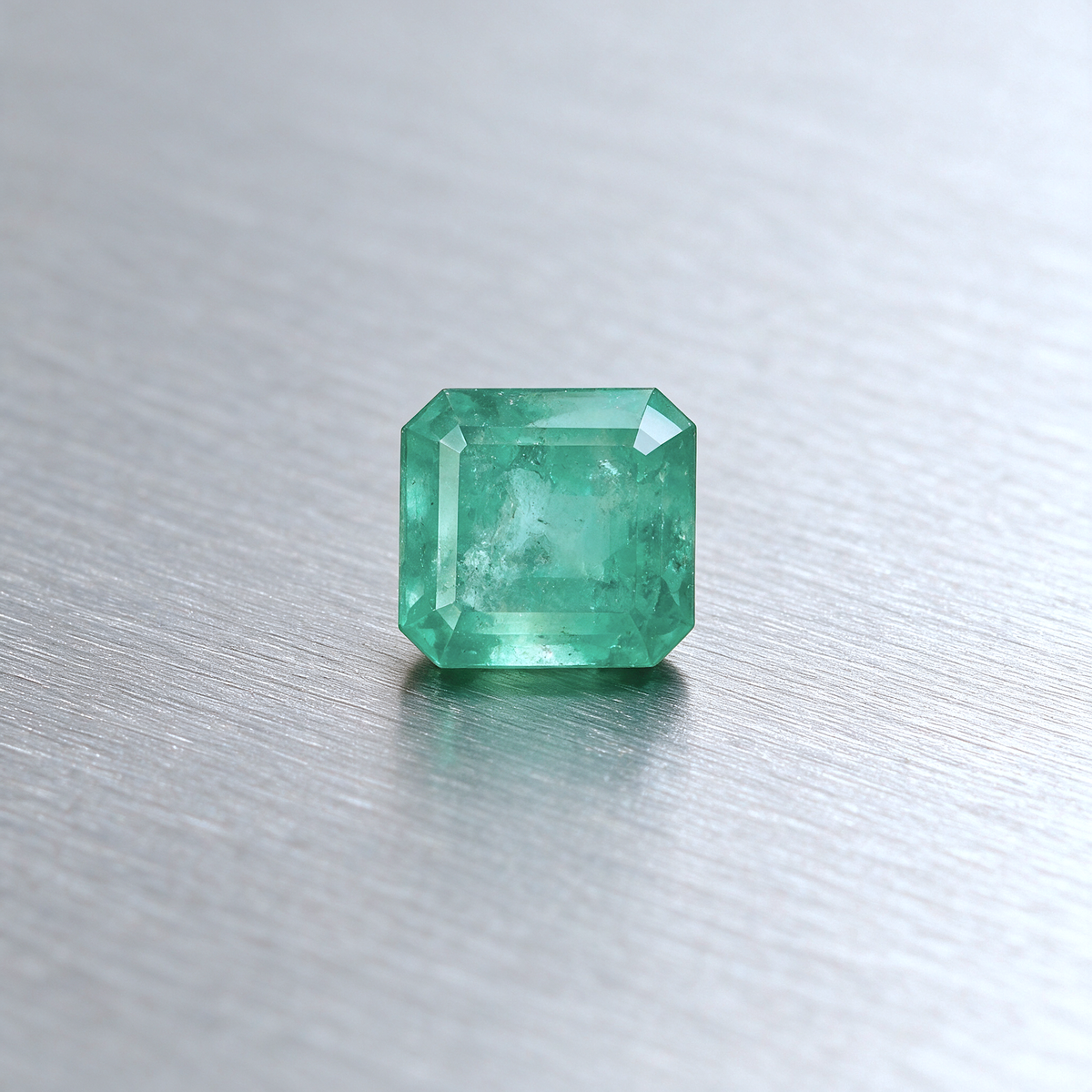 A single emerald-cut green emerald with visible internal inclusions, resting on a brushed silver metal surface.