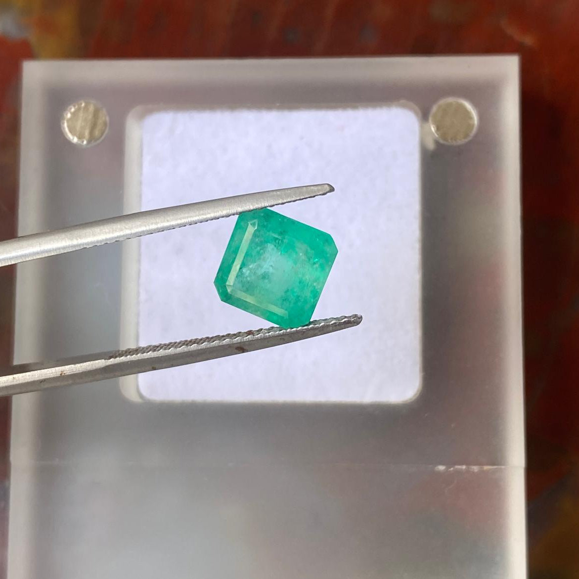 A green gemstone held by metal tweezers over a translucent white square display block, with a warm brown textured background.
