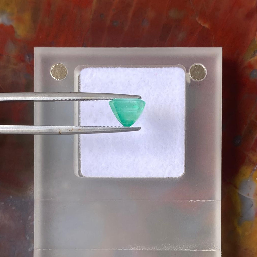 A small green faceted gemstone held by metal tweezers above a clear acrylic mounting block, photographed against a mottled warm brown and orange surface.