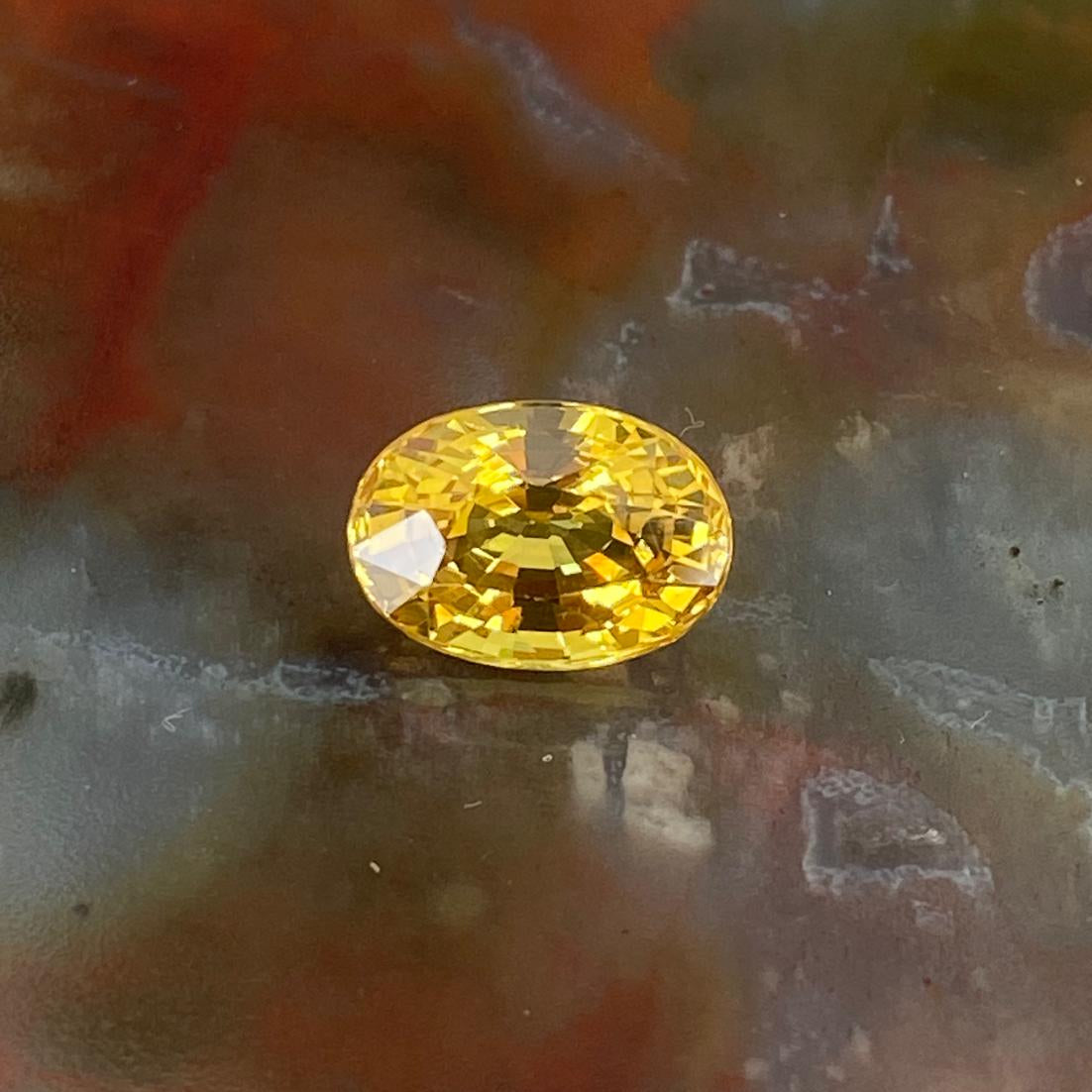 An oval-cut yellow sapphire with a faceted finish, resting on a textured, mottled surface of brown, gray, and red tones.