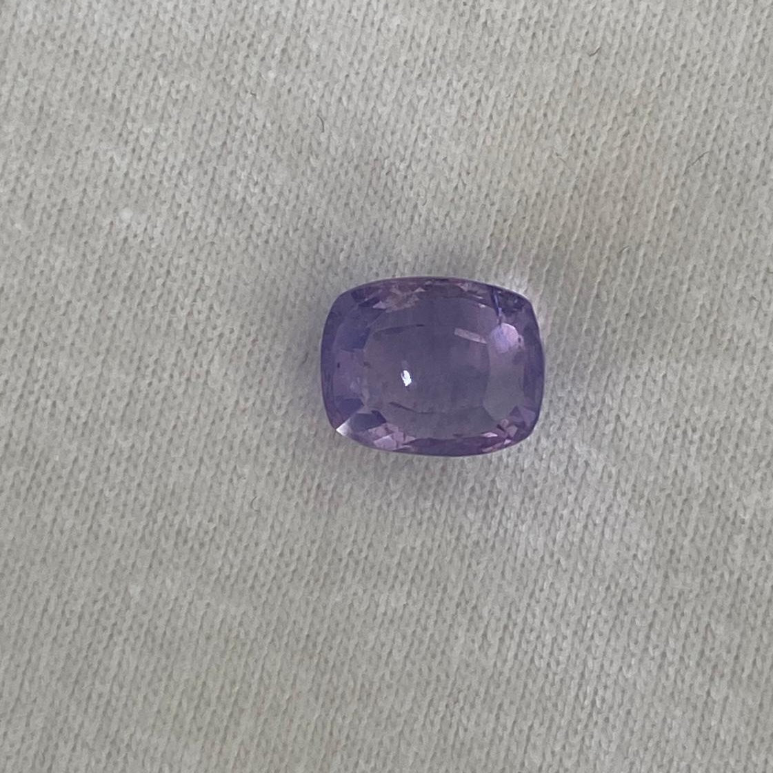A single cushion-cut purple sapphire, resting on a textured light gray fabric background.