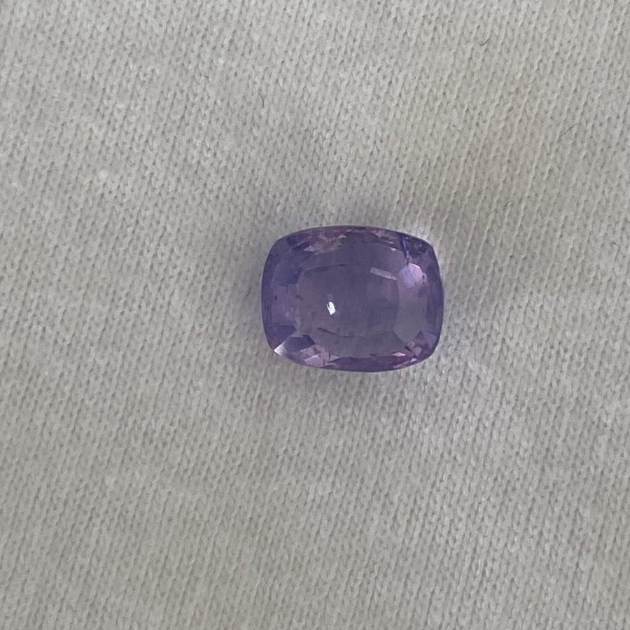 A single cushion-cut purple sapphire, resting on a textured light gray fabric background.