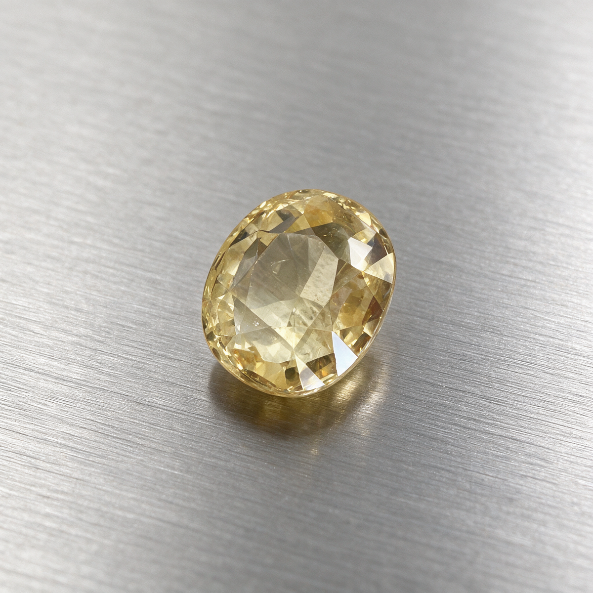A single oval-cut yellow sapphire with a faceted surface, resting on a brushed silver metal background.