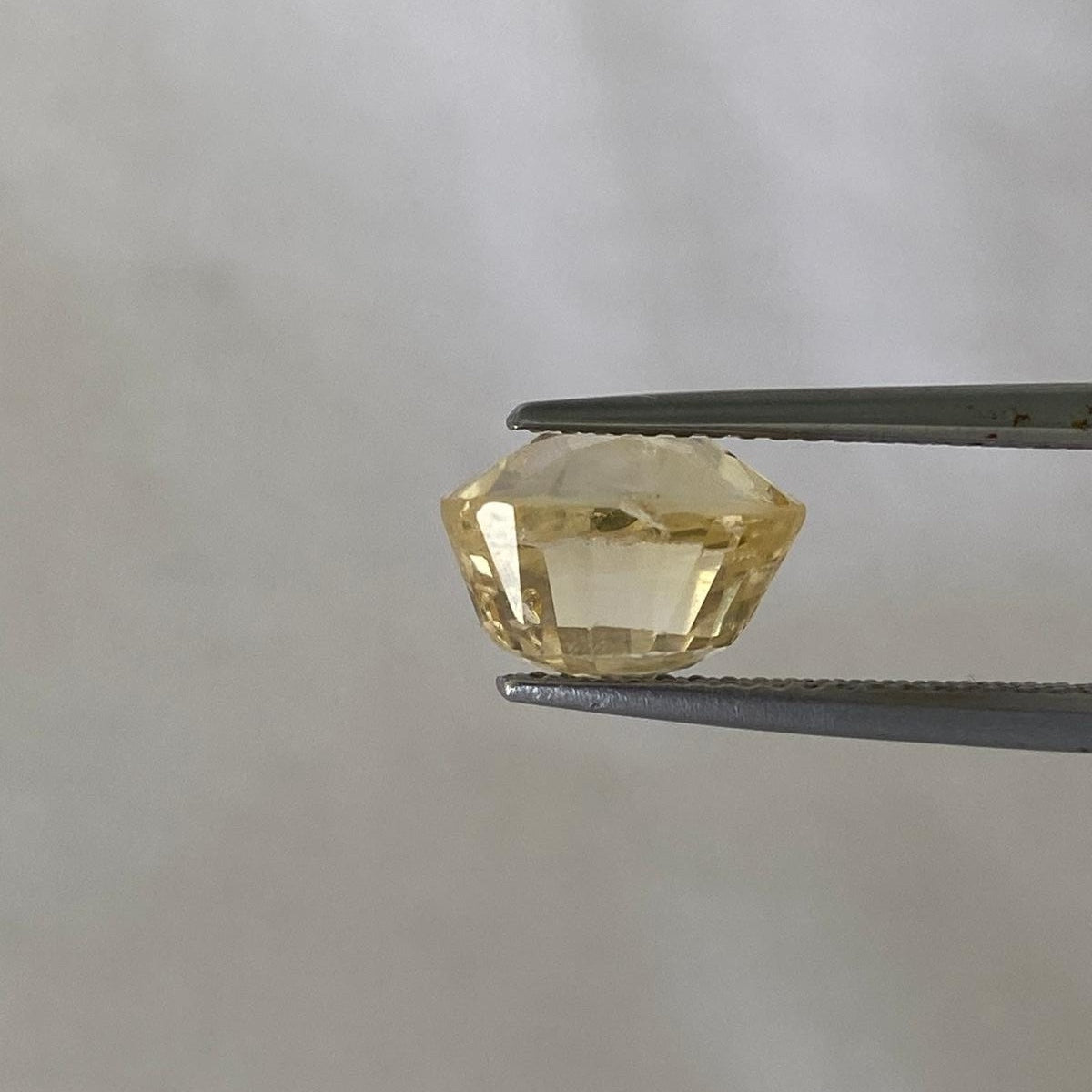 A faceted yellow sapphire held between metal tweezers against a plain light gray background.